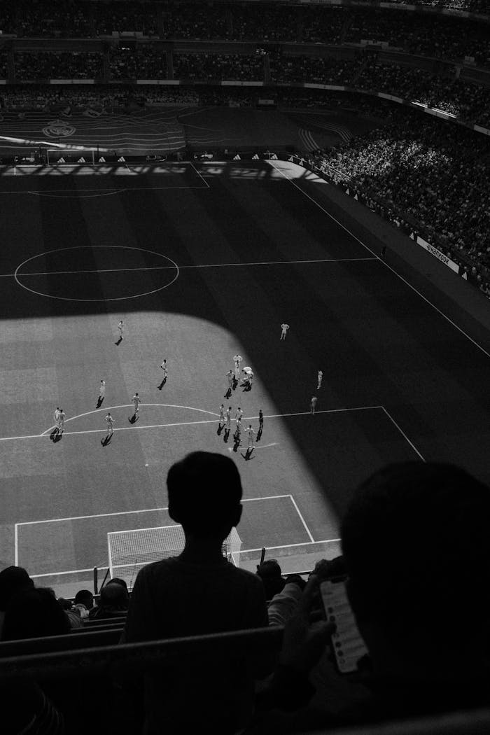 Diensten Aerial view of a football match with spectators silhouetted in the foreground, captured in striking black and white.